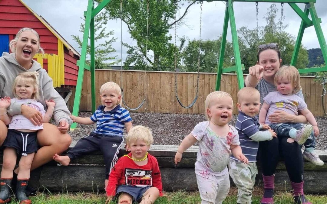 Parents and children sit in front of a sand pit and swing set