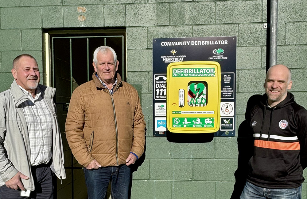 Three men stand beside a bright yellow AED cabinet labeled 'Community Defibrillator' mounted on a green wall outside. 2) Outdoor scene with three men next to a public defibrillator on a wall. 3) Three adults posing near a defibrillator mounted on a green brick wall. 4) Public access AED defibrillator unit on a wall with three men nearby. 5) AED defibrillator cabinet and three men standing outdoors.