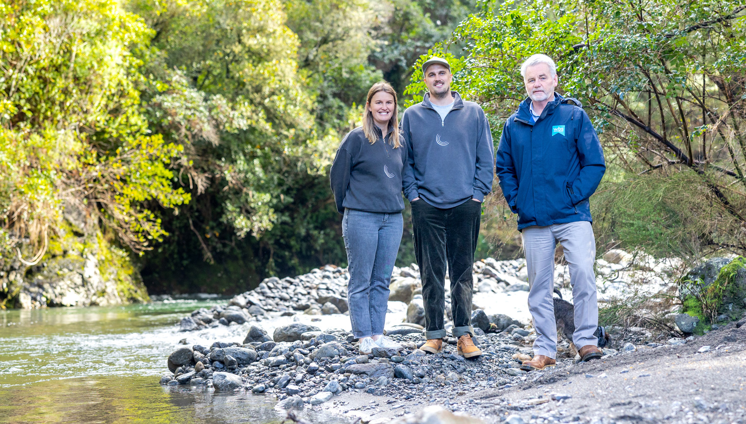 Three people stand together on a rocky riverbank in a green forest, posing for a photo.