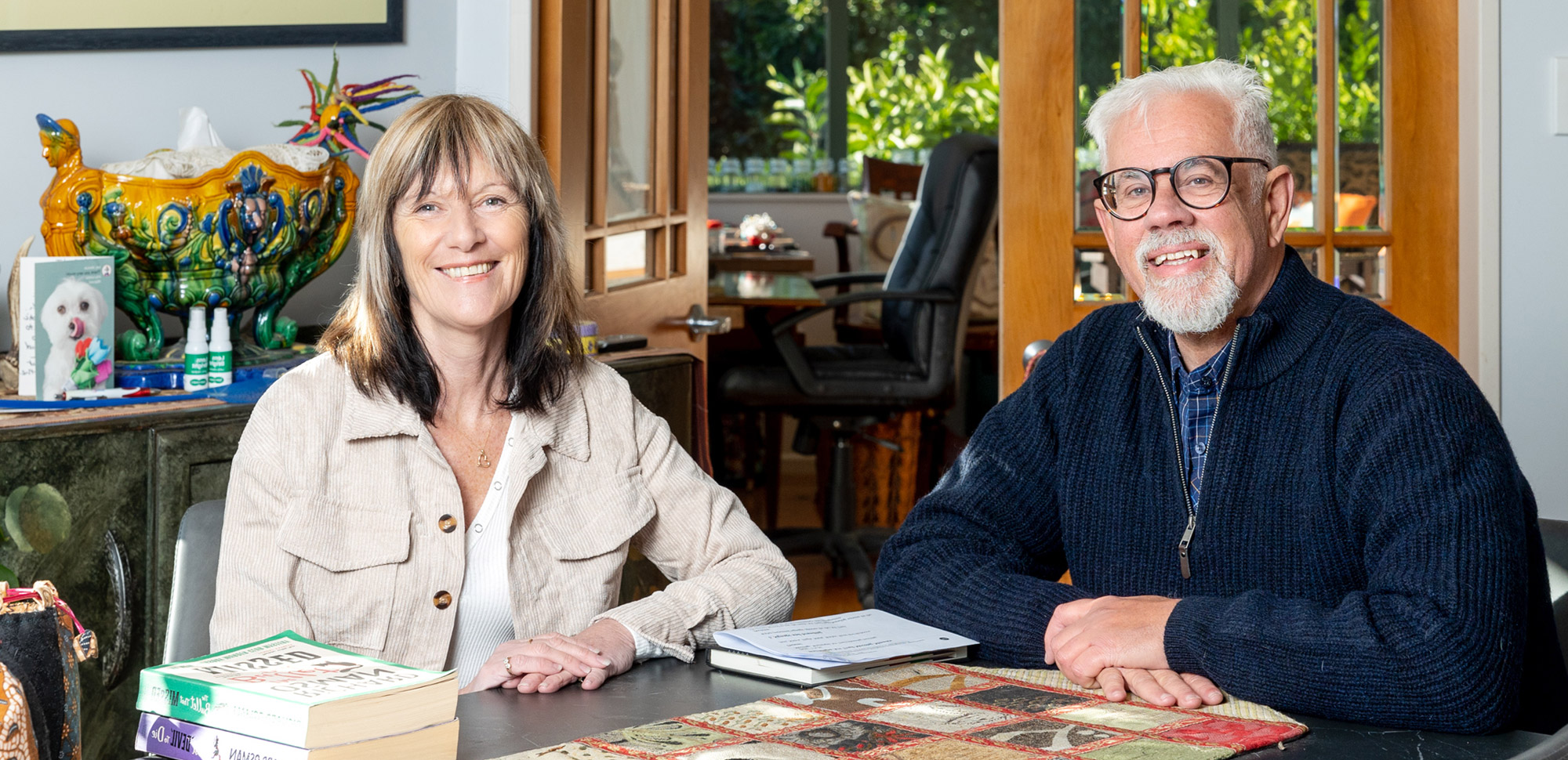 Two adults sit at a table with papers and notebooks, smiling at the camera in a bright room.