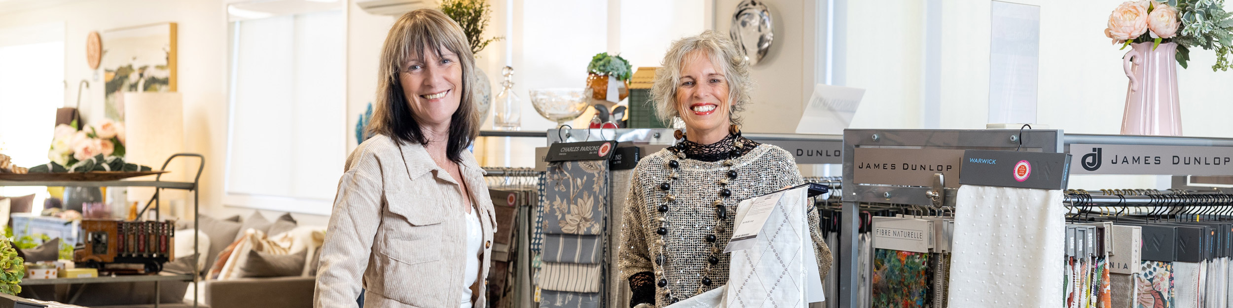 Two smiling women stand among fabric racks and decor in a bright showroom.