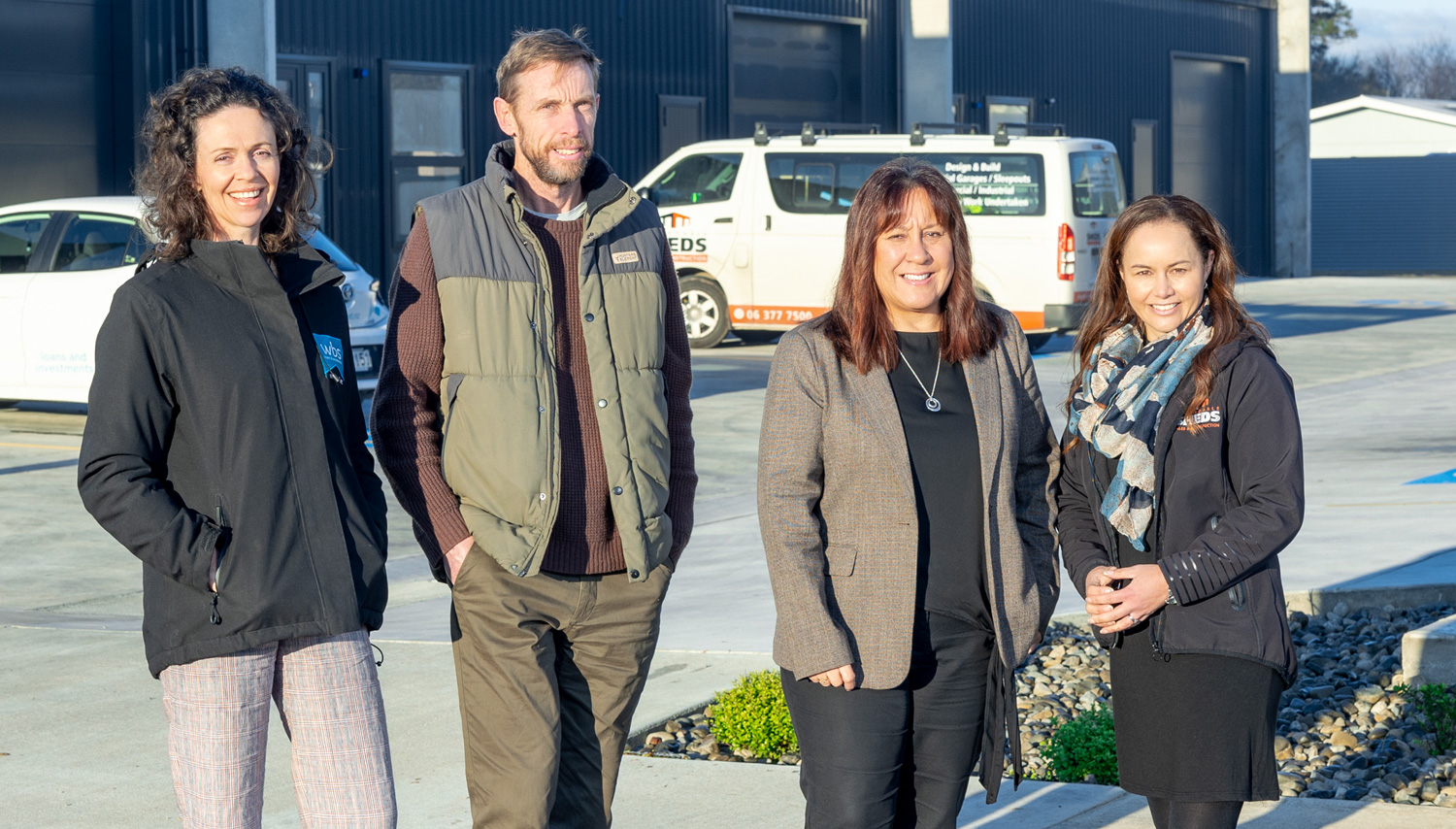 Four adults stand together outdoors in front of a blue industrial building and white service vans, smiling at the camera.