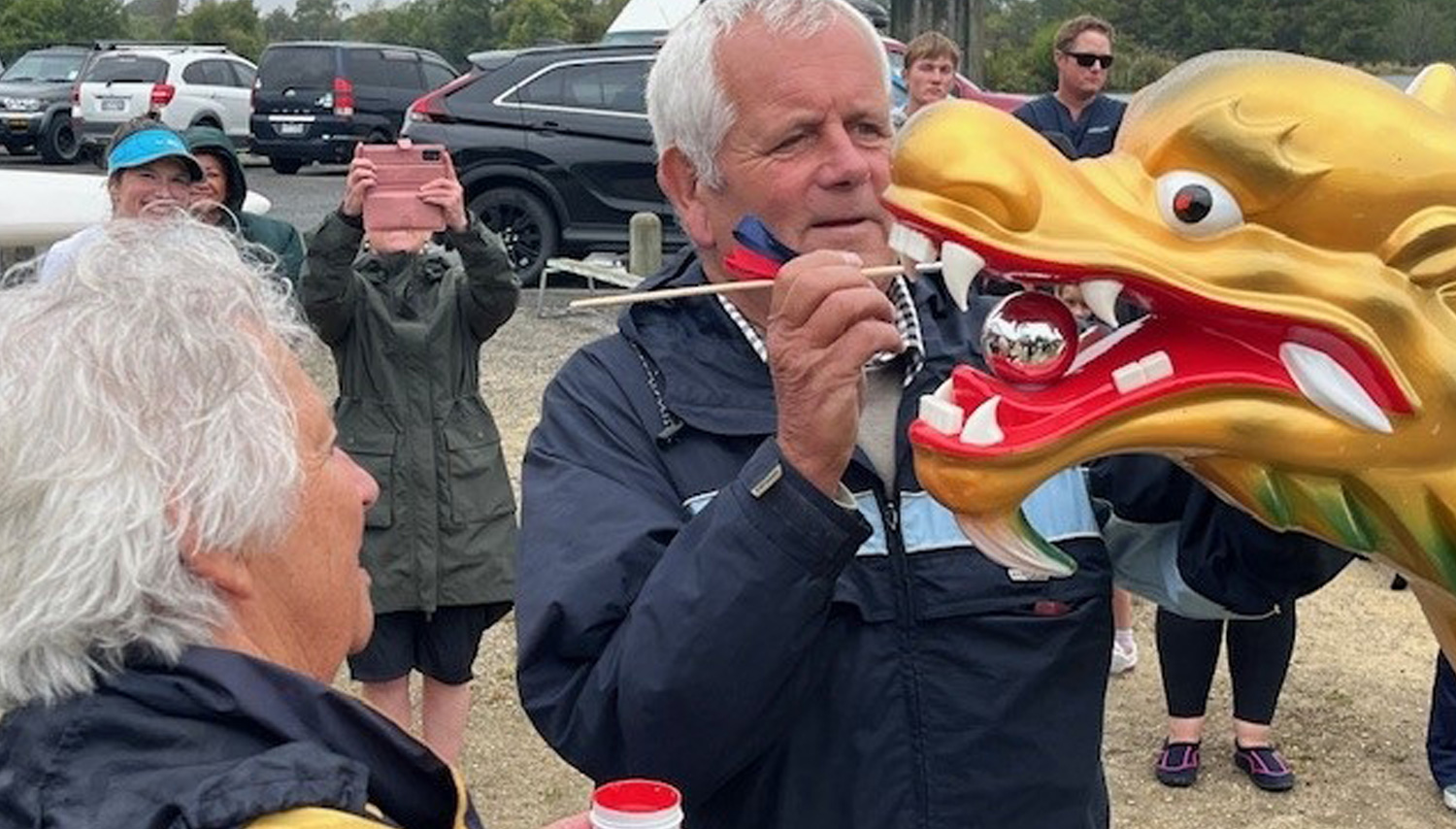 An older man holding a dragon head prop near his face at an outdoor event, others watch and take photos in a parking lot.