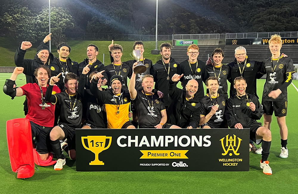 Wellington Hockey players in black celebrate their Premier One championship, raising the trophy with a CHAMPIONS banner in the foreground.